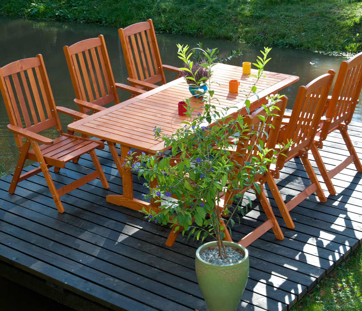 Wooden outdoor dining set with chairs on a deck by water, potted plant in foreground